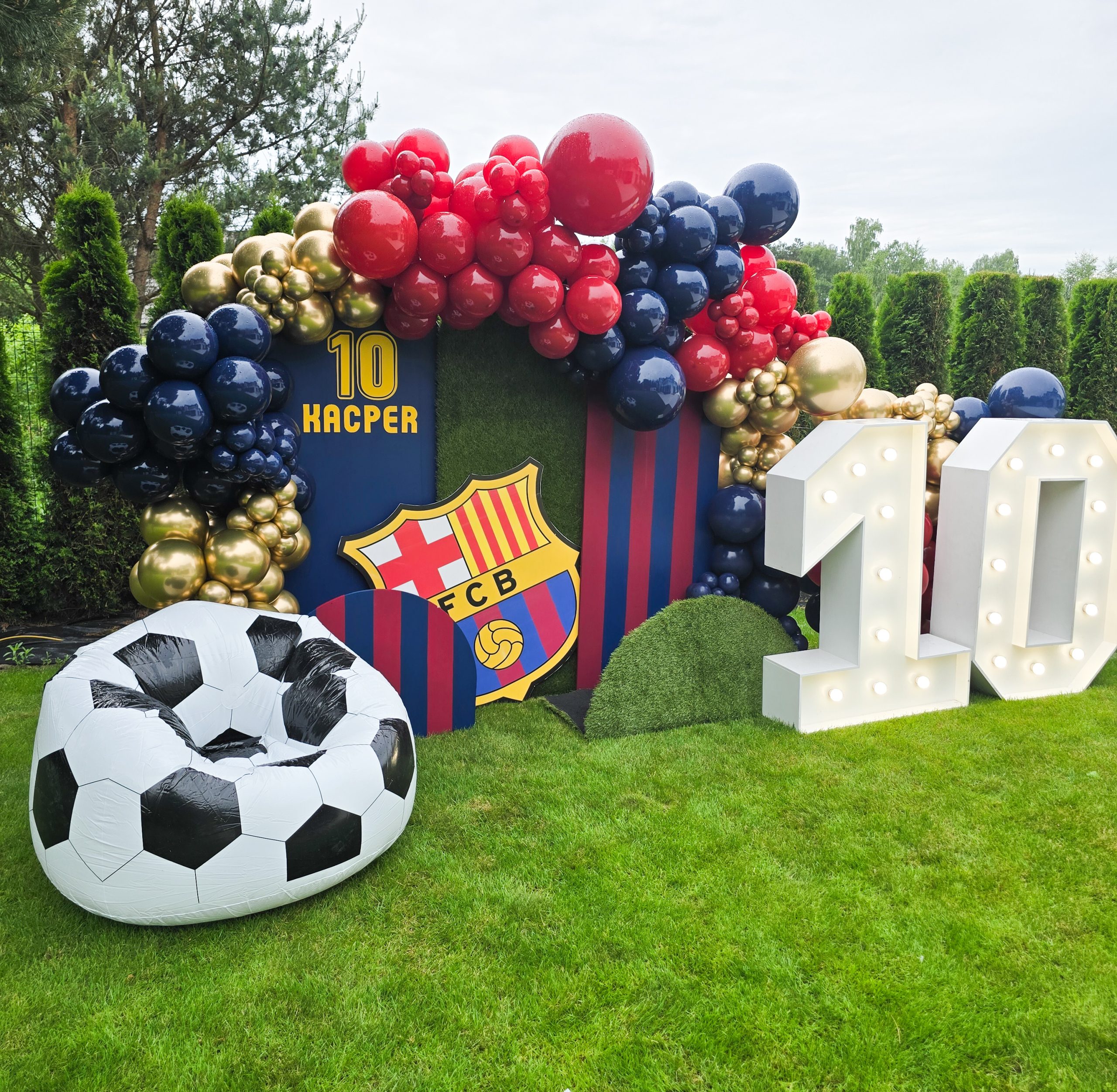 Dekoracja urodzinowa w tematyce piłkarskiej klubu FC Barcelona ustawiona na trawniku. Zawiera dużą girlandę z balonów w kolorach czerwonym, granatowym i złotym, tło z herbem klubu oraz imieniem „KACPER” i numerem „10”. Po bokach znajdują się duże, podświetlane cyfry „10” oraz pufa w kształcie piłki nożnej.