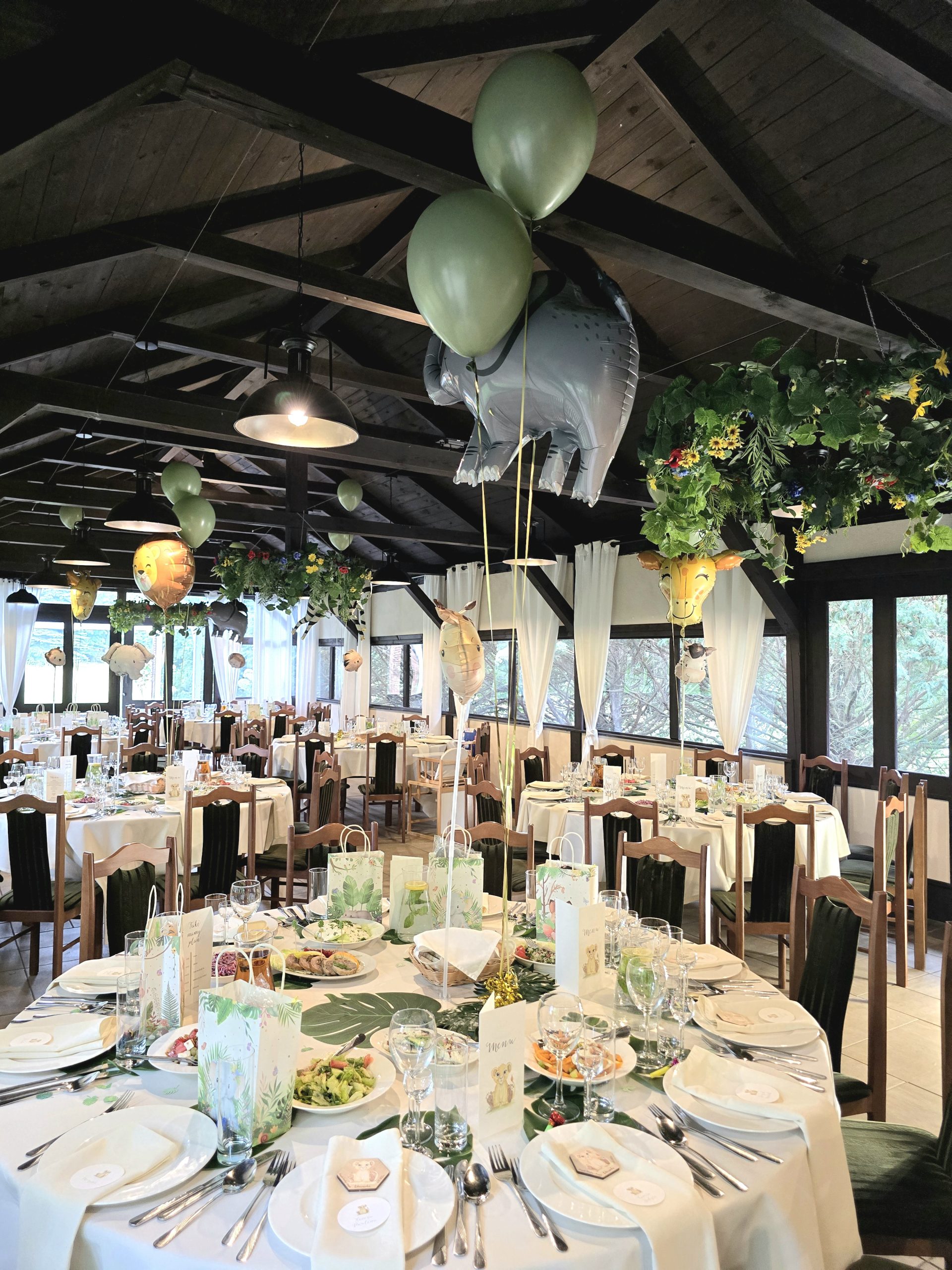 Sala restauracyjna z drewnianym sufitem przygotowana na przyjęcie okolicznościowe. Okrągłe stoły są nakryte białymi obrusami i zastawą. Nad stołami unoszą się balony z motywem safari, w tym duży balon w kształcie szarego słonia.