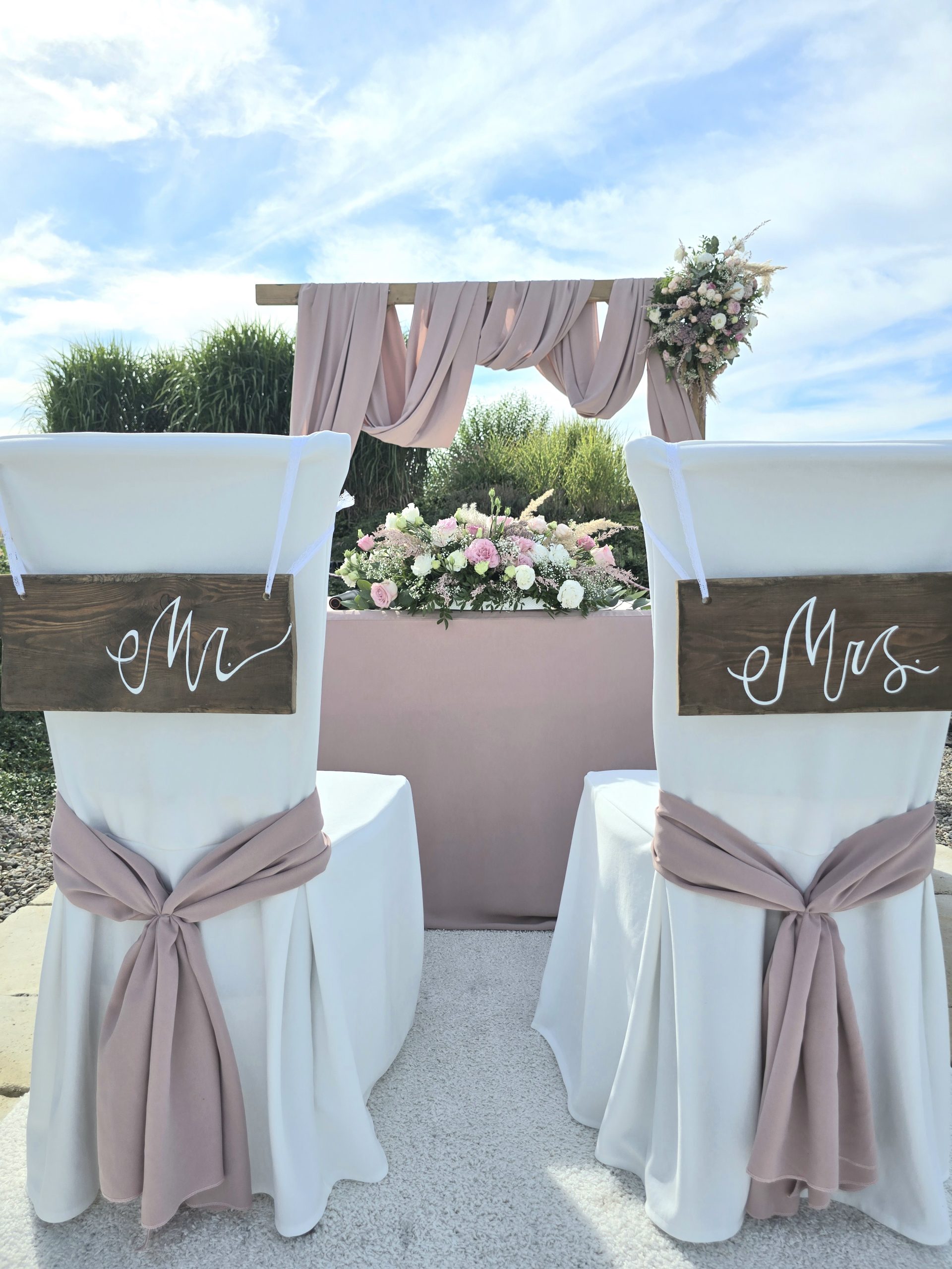 Widok z tyłu na dwa białe krzesła przygotowane dla pary młodej podczas ceremonii ślubnej w plenerze. Na oparciach wiszą drewniane tabliczki z napisami "Mr" oraz "Mrs". Krzesła są udekorowane różową tkaniną, a w tle widać ołtarz z kwiatami pod błękitnym niebem.